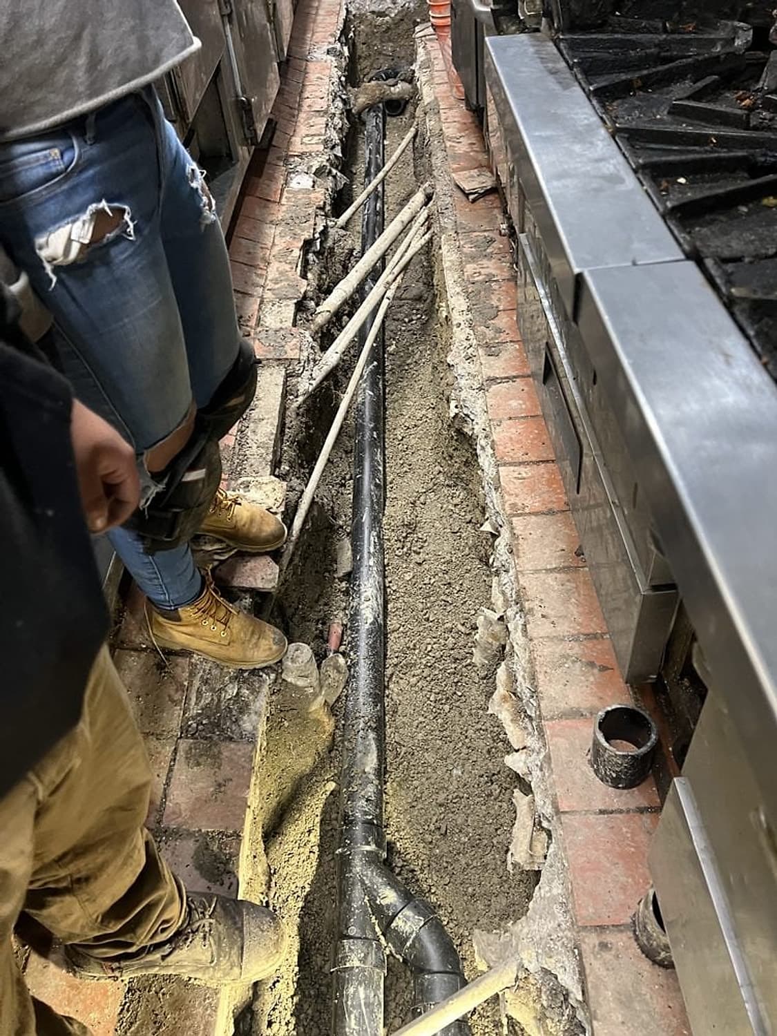 Open trench in a commercial kitchen floor with a new black ABS drain line running beside the cooking equipment