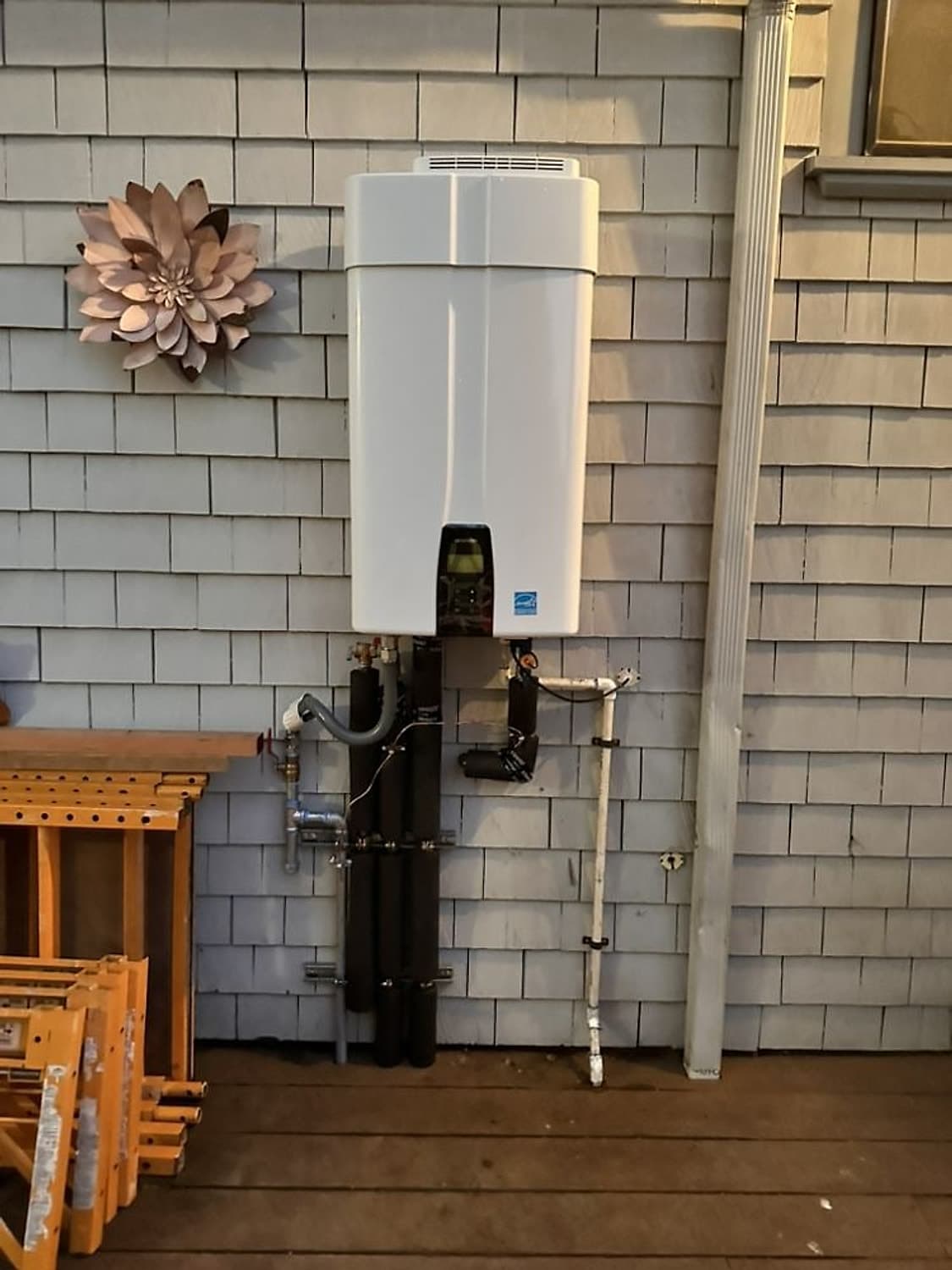 White tankless water heater mounted on a gray shingle exterior wall with insulated supply lines below
